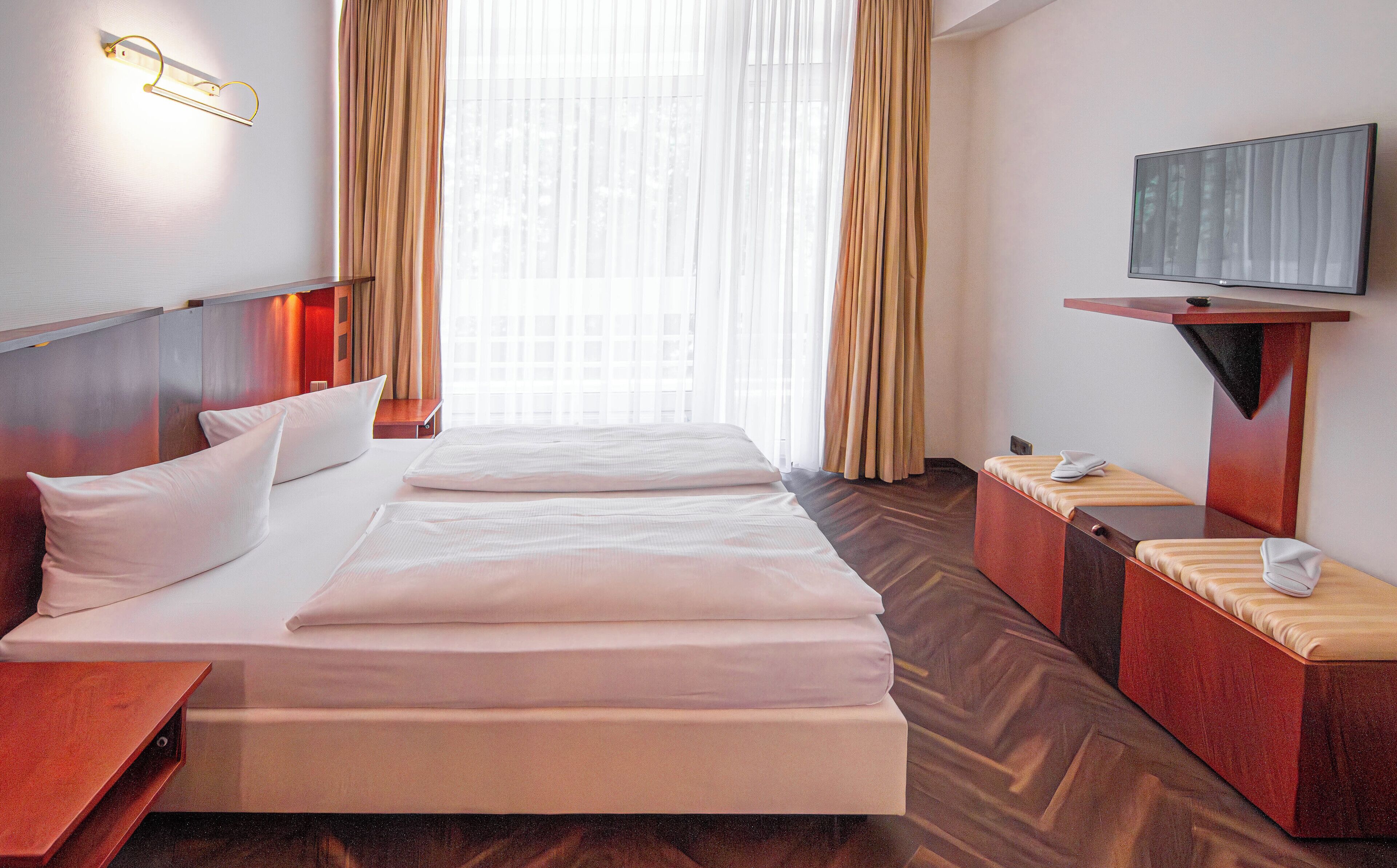 A hotel room with a bed, a TV, a desk, and a window with curtains.