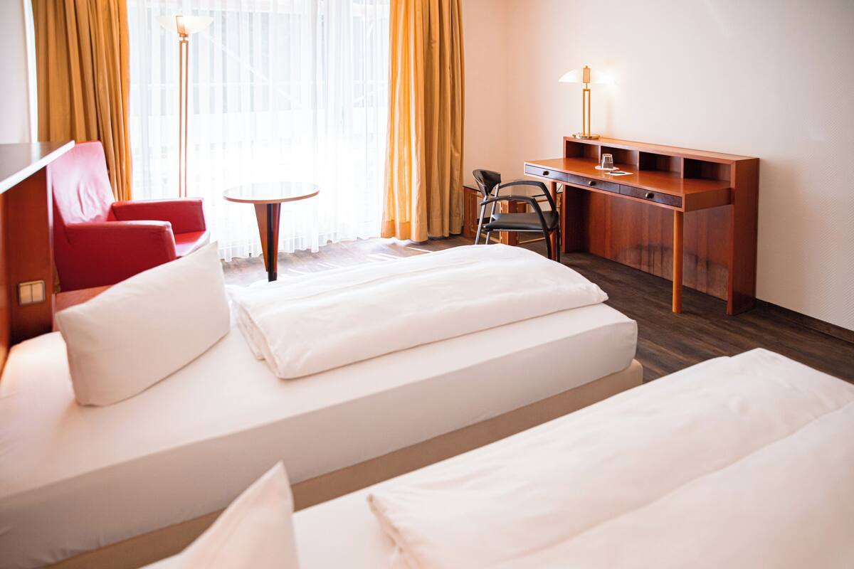 A hotel room with two beds, a desk, a chair, and a red armchair.