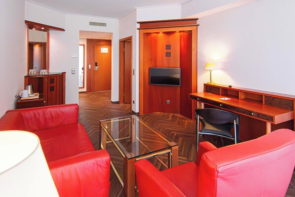 A hotel room with a glass-top coffee table, red leather sofa, a wooden desk with a flat-screen TV, and a lamp.