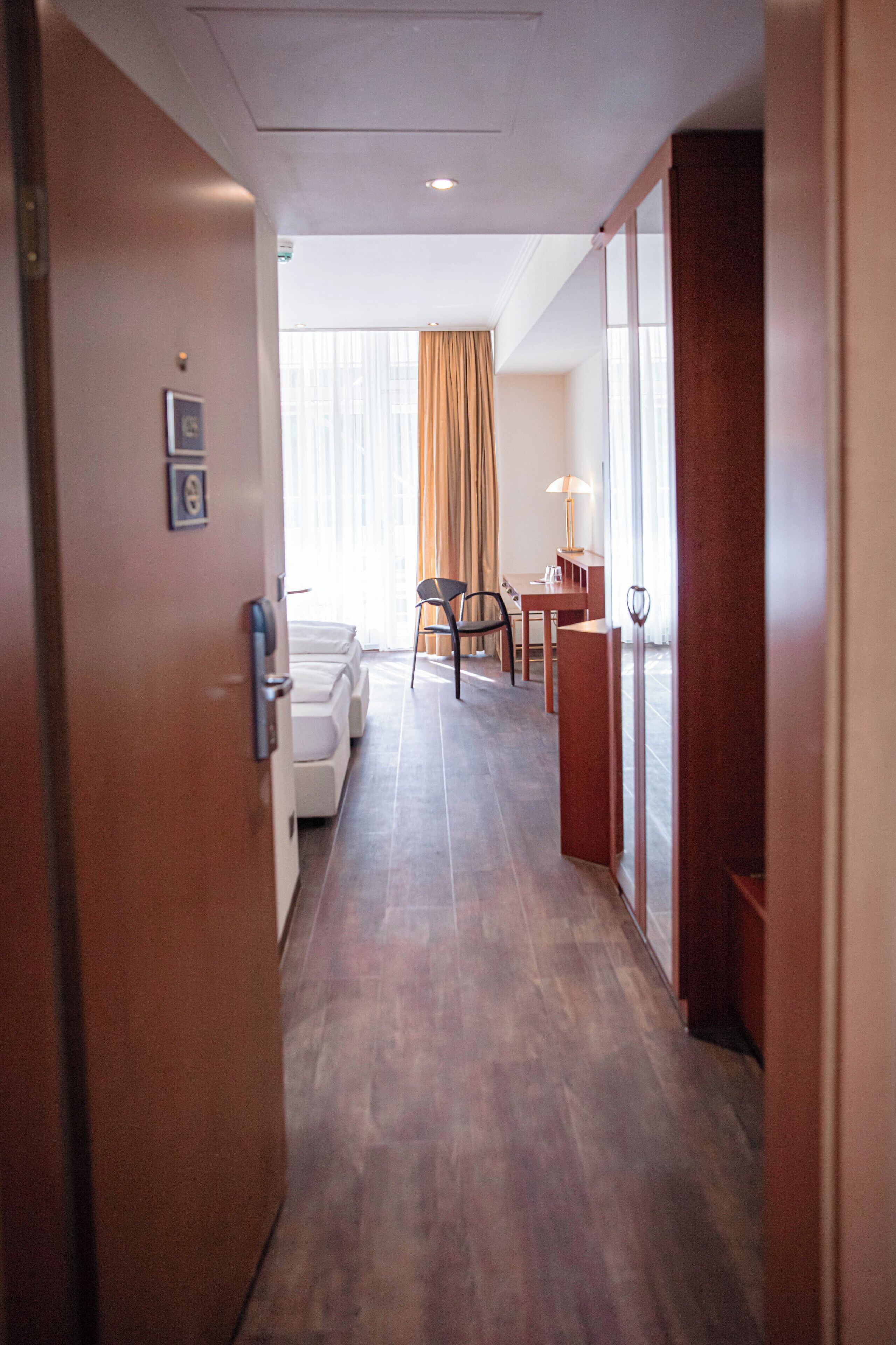 A hotel corridor with a wooden floor, a seating area with a table and chairs, and a view of a room with a window and curtains.