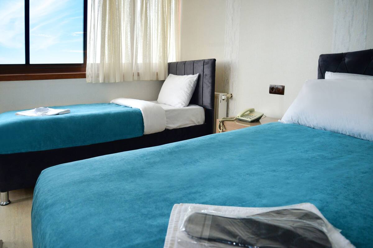 A hotel room with two beds, each with a blue bedspread and white pillows. There is a window with sheer curtains, a bedside table with a telephone, and a wall socket.