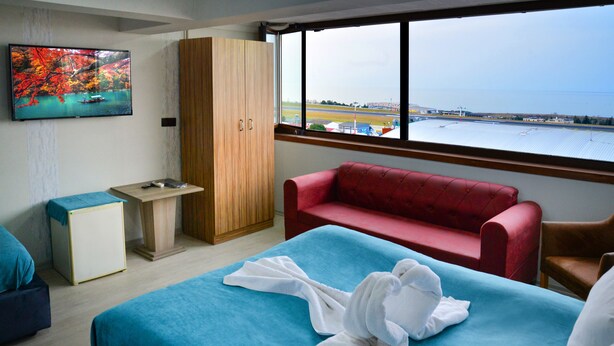 A hotel room with a bed, a red sofa, a wooden wardrobe, a television, and a window with a view of a body of water and airport. - Elite Holiday Hotel (Trabzon)