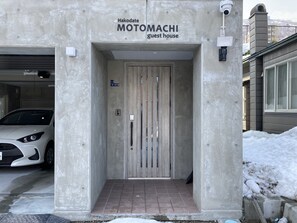 Guests enter the Hakodate MOTOMACHI guest house through a modern entrance with a light wood door and a sign above the door.