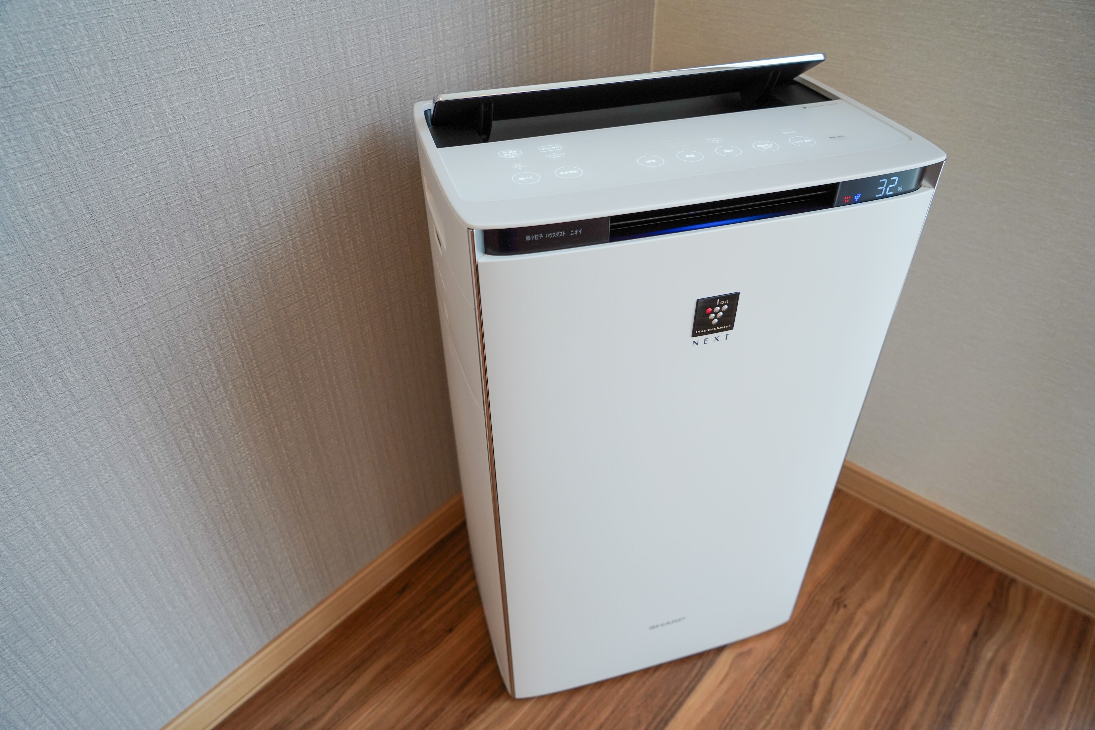 A white, compact, wall-mounted air conditioning unit with a digital display, located in a corner with wooden flooring and light-colored walls.