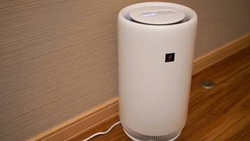 A white, cylindrical air purifier with a control panel and a small logo, placed on a wooden floor against a textured wall.