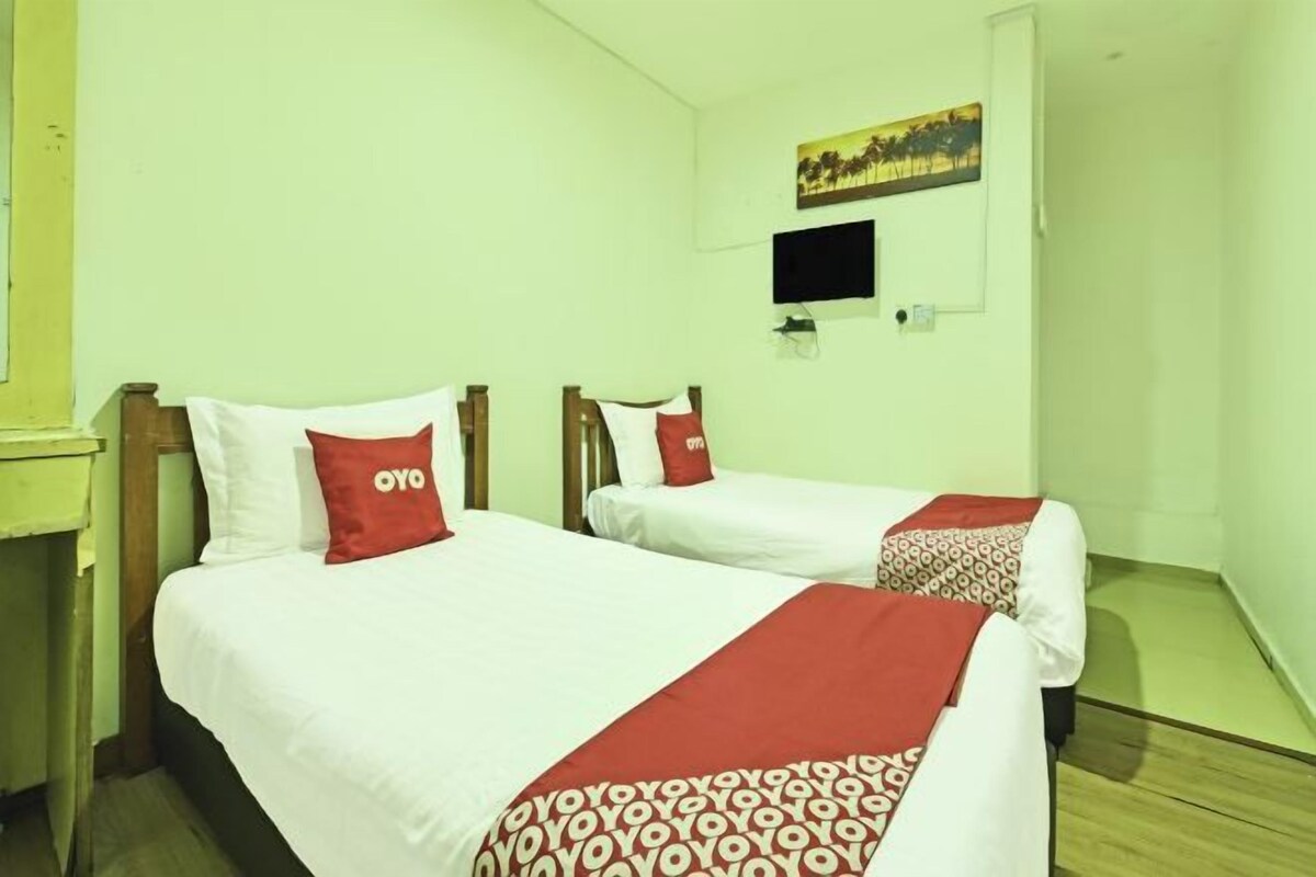 A hotel room with two beds, each with white bedding and red accents, a flat-screen TV mounted on the wall, and a painting above the TV.