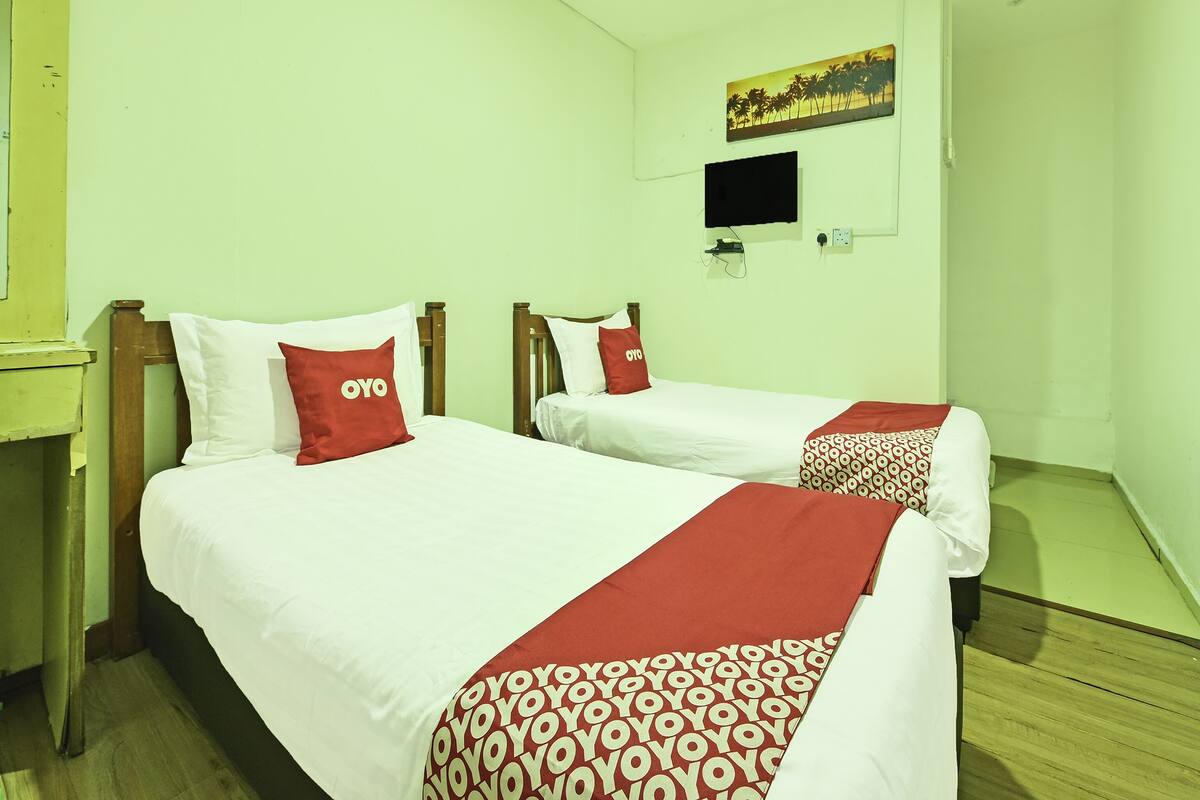 Habitación de hotel con dos camas, cada una con ropa blanca y almohadas rojas. Hay un televisor montado en la pared y un cuadro encima.