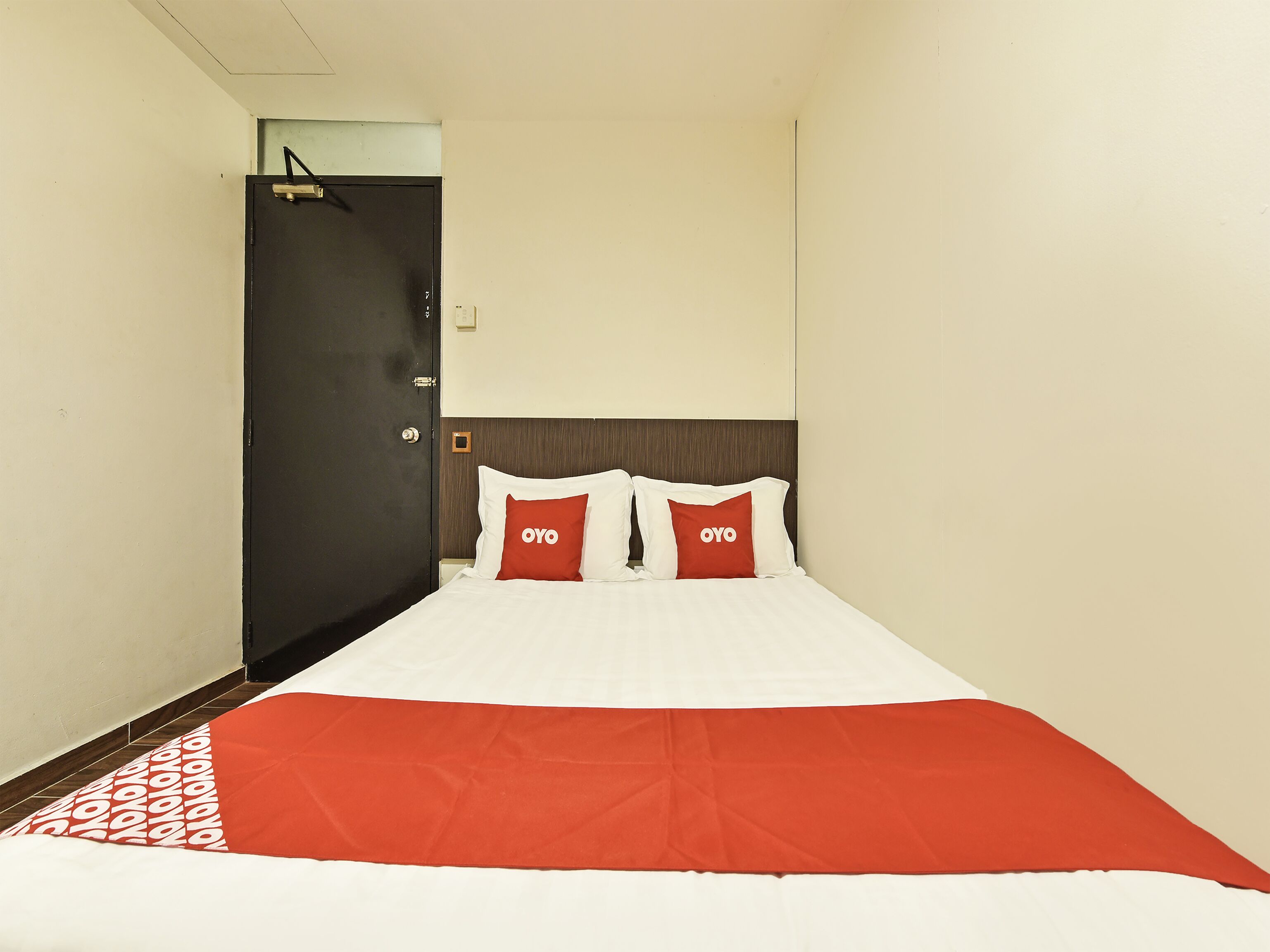 A hotel room with a bed, two pillows with the logo "OYO", a dark door, and a light-colored wall.
