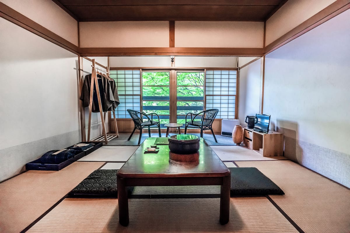 Een traditionele Japanse kamer met tatami vloeren, een lage tafel met een groene kom, twee stoelen en een houten kast met een laptop.