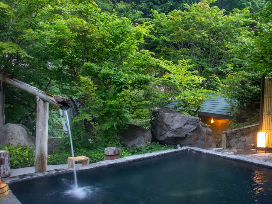 A hot spring surrounded by lush greenery and a wooden structure with a light.