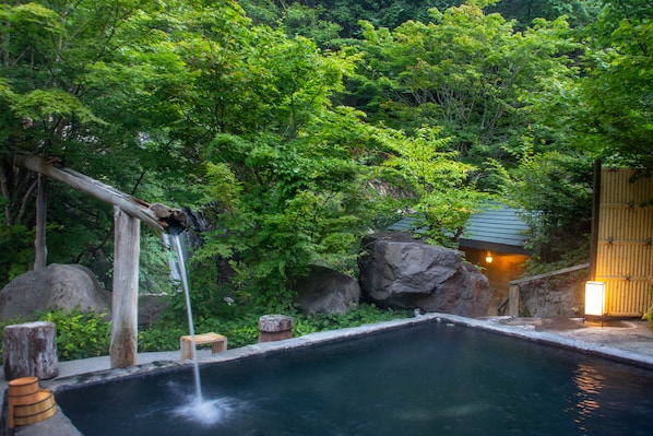 A hot spring surrounded by lush greenery and a wooden structure with a light.