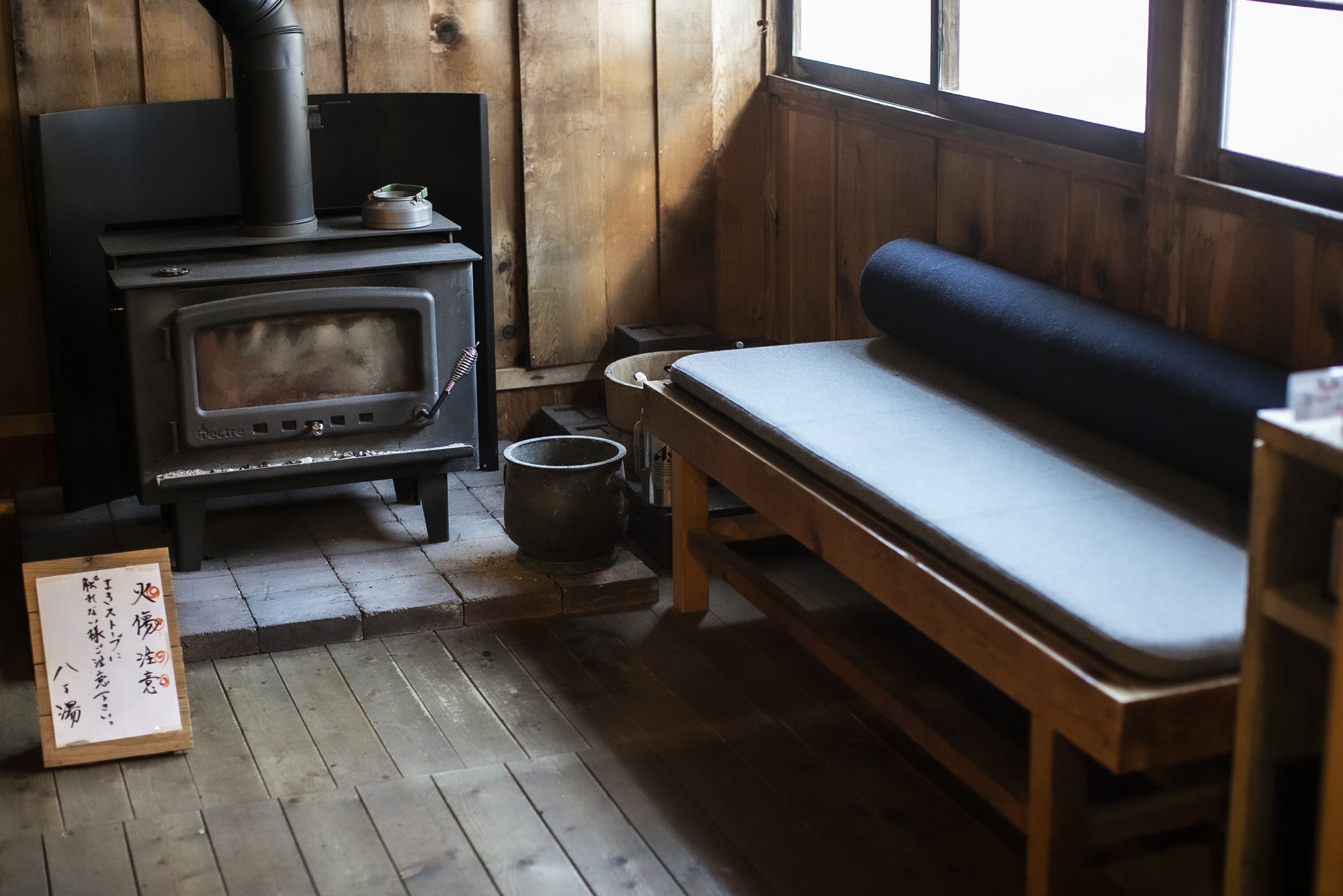 A wooden interior with a wood-burning stove, a bench, and a sign with Japanese characters.
