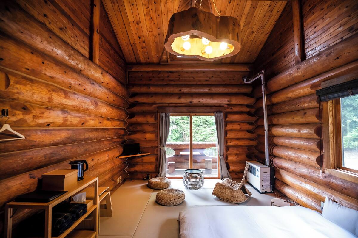 Een blokhutkamer met een bed, nachtkastje, hanglamp en uitzicht op de natuur door een raam.