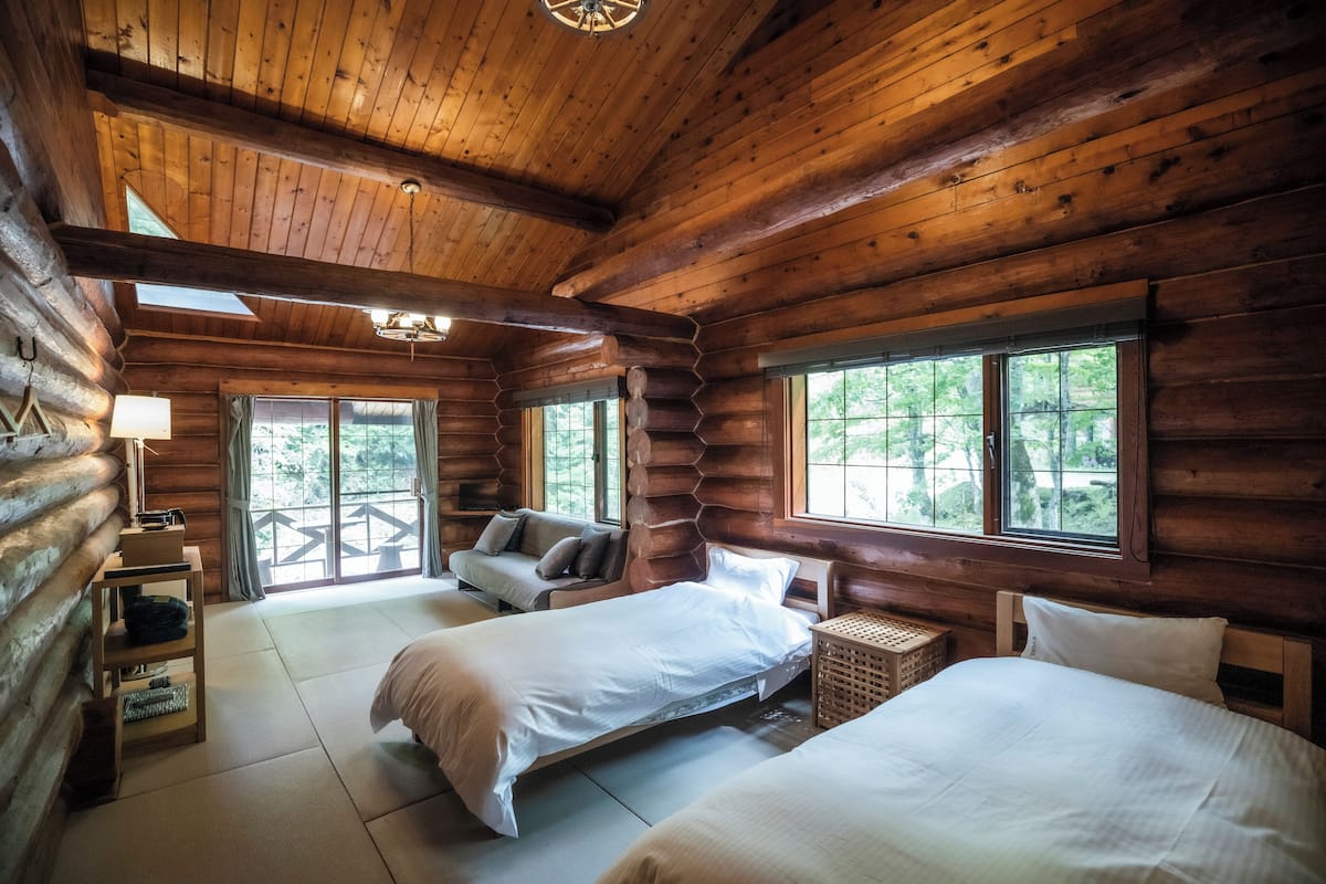 Een slaapkamer in een blokhut met twee bedden, een sofa en een raam met uitzicht op groen.