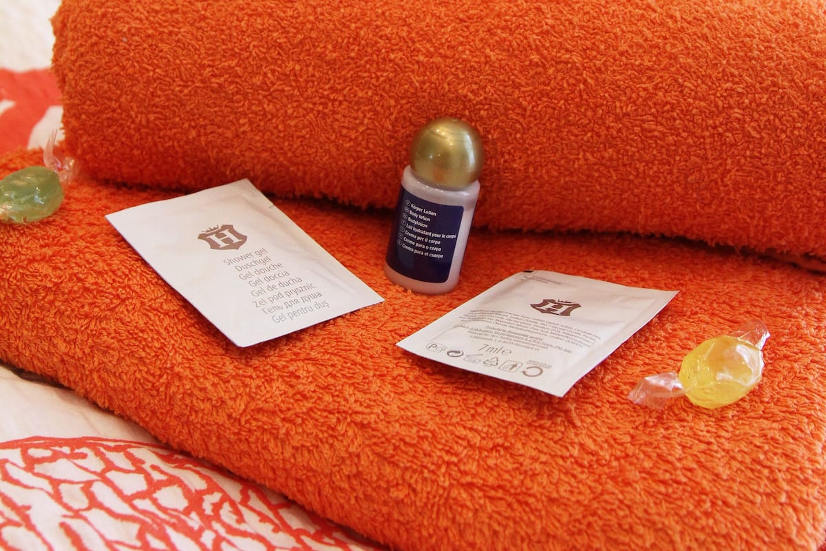 A neatly made bed with an orange towel and a bottle of lotion.