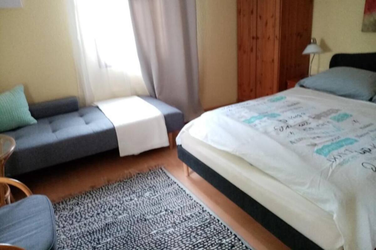 A bedroom with a bed, a sofa, a small table, and a window with curtains.