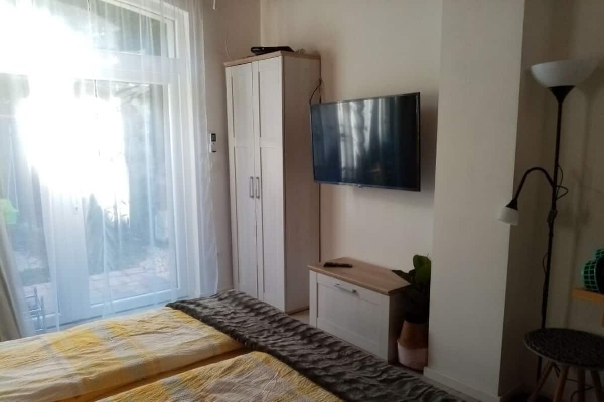 A bedroom with a bed, a TV mounted on the wall, a wardrobe, and a desk with a plant.