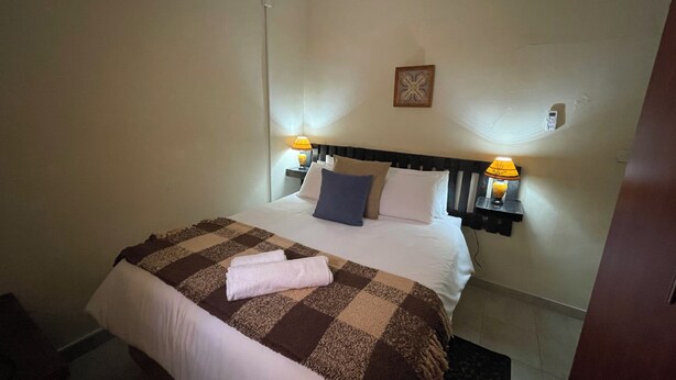 A hotel room with a bed, two bedside lamps, a wall-mounted air conditioner, and a small decorative item on the wall. - Abrama Bed And Breakfast (Francistown)