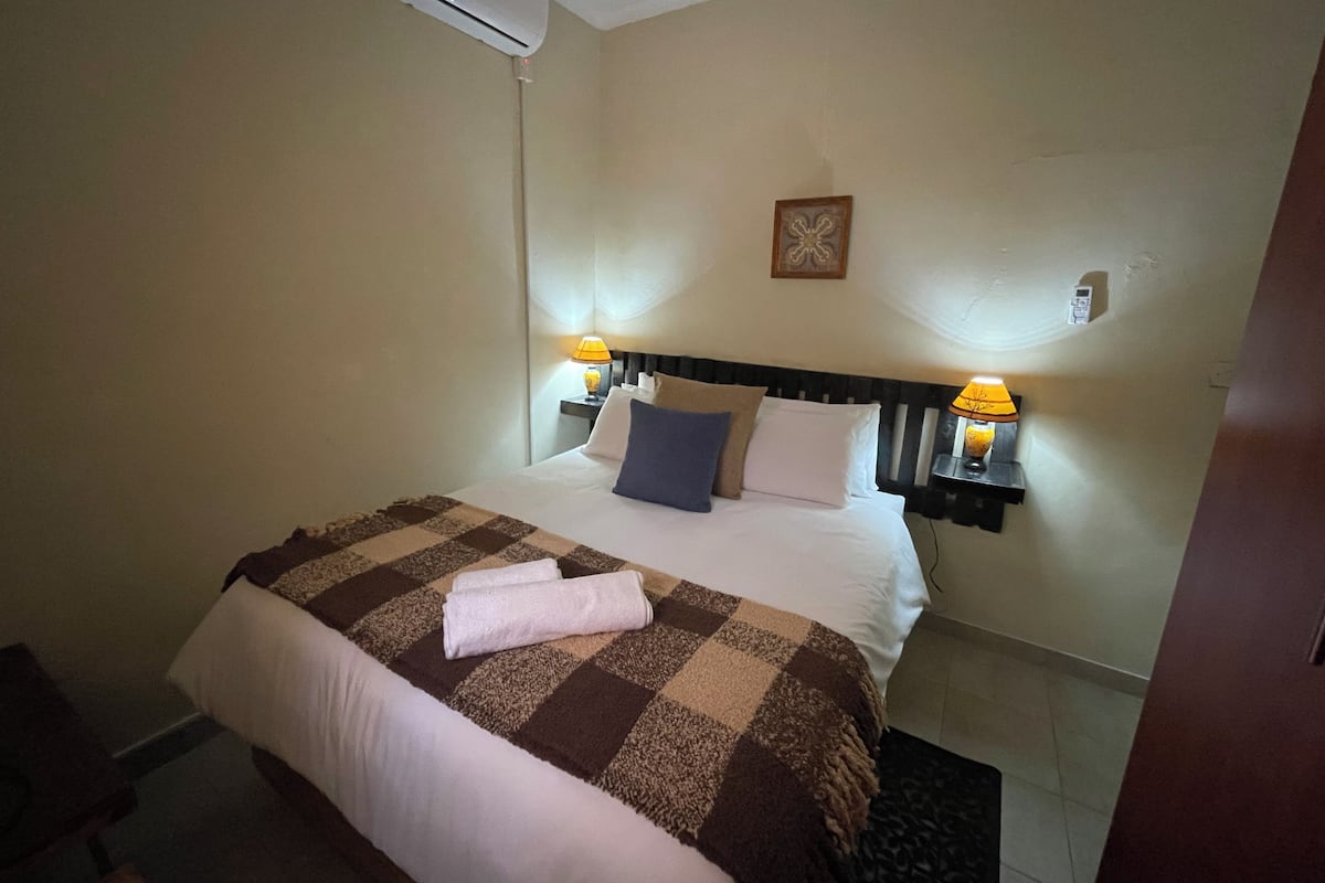 Habitación de hotel con cama, dos lámparas de noche, aire acondicionado de pared y un pequeño adorno en la pared.