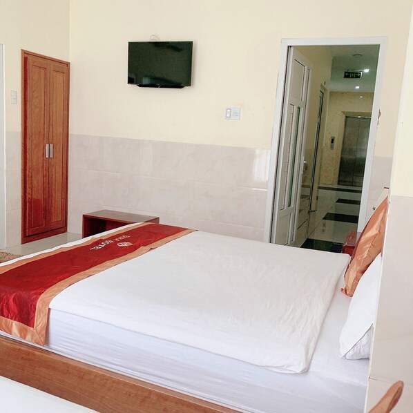 A hotel room with a bed, a wooden wardrobe, a television mounted on the wall, and a door leading to a hallway.