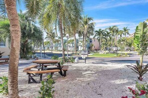 A paved outdoor area with picnic tables, palm trees, and a building complex in the background.