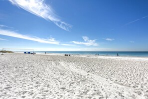 A wide sandy beach with footprints and tire tracks, a clear blue sky, and a calm sea.