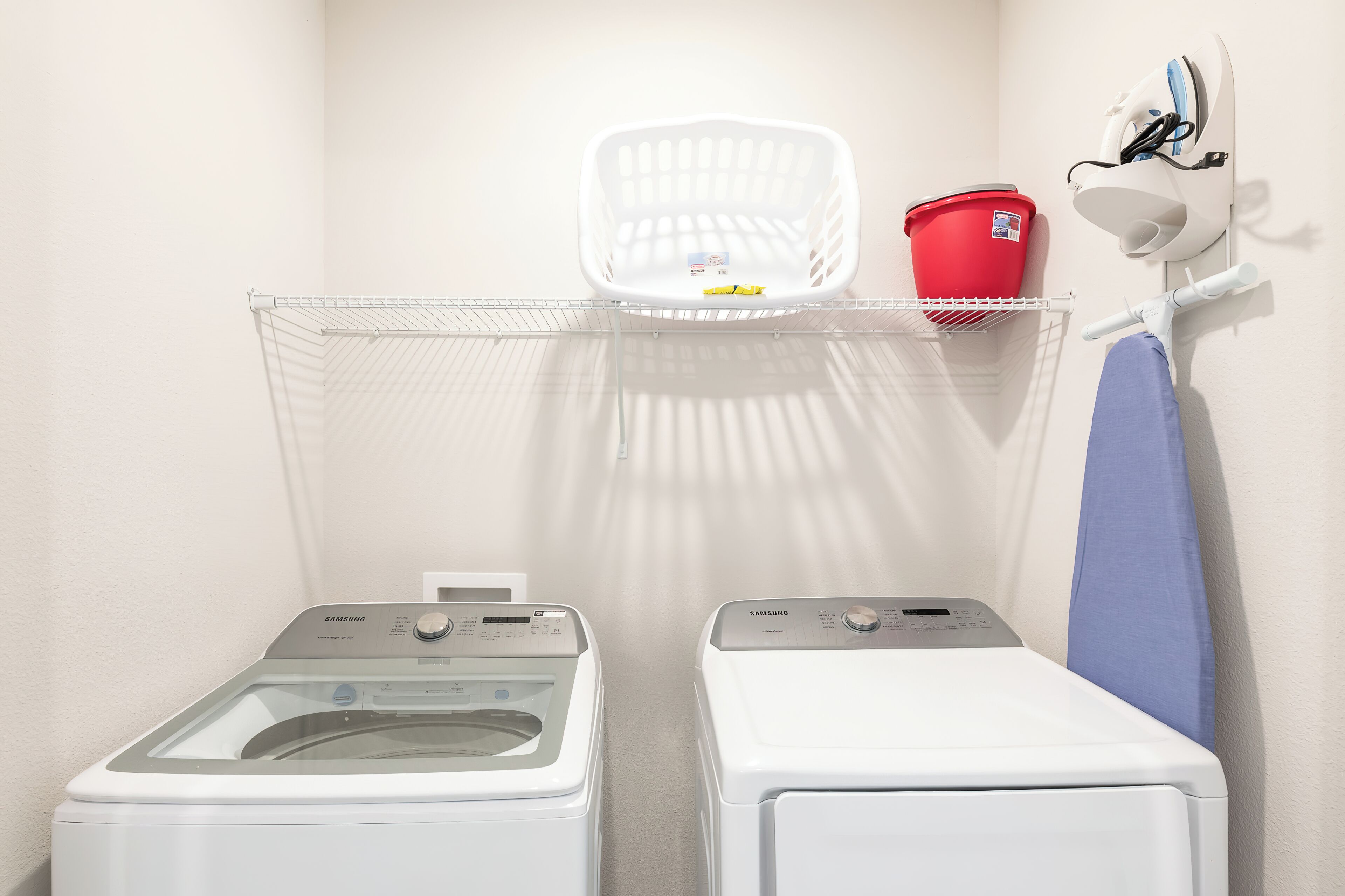 A laundry area with two front-loading washing machines, a white laundry basket, a red bucket, and a wall-mounted iron.