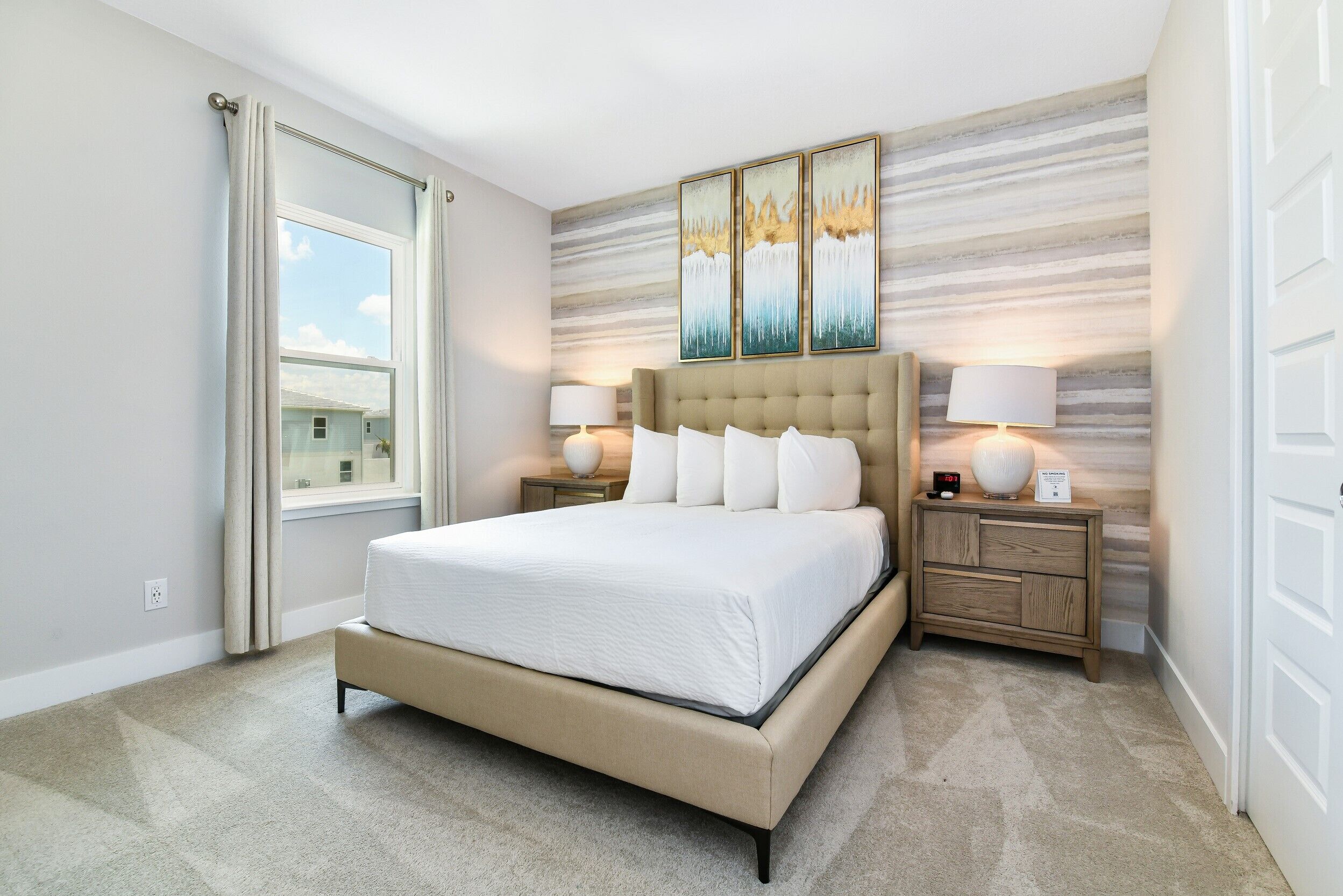 A bedroom with a large bed, two bedside tables with lamps, a window with curtains, and a wall with three framed artworks.