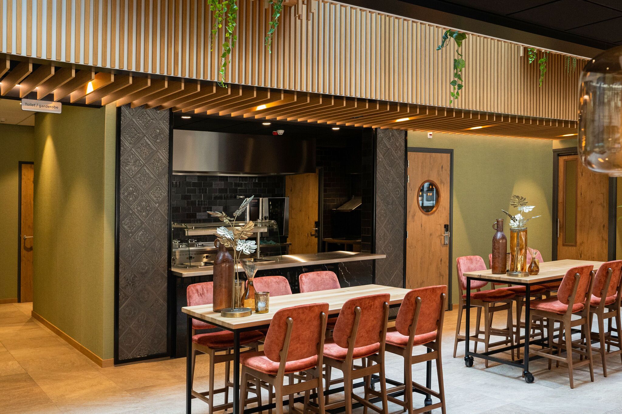 A restaurant with a long wooden table and red upholstered chairs, a kitchen area, and a sign indicating the location of the toilet and garden room.