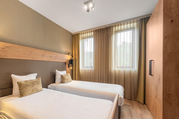 Ein Hotelzimmer mit zwei Betten, einem Holzkopfende, einem Nachttisch mit Lampe und einem großen Fenster mit Vorhängen.