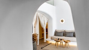 A modern interior with a white sofa, two wooden side tables, a patterned rug, and a macramé curtain. - Victoria Caves (Santorini)