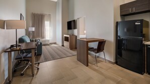 Ein modernes Hotelzimmer mit Küchenzeile, Schreibtisch, Stuhl, Fernseher und einem großen Fenster mit Vorhängen.