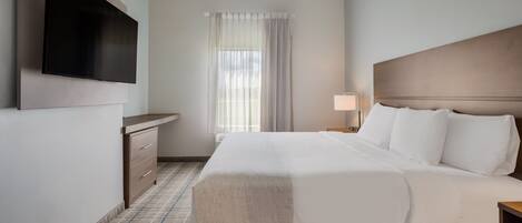 Una habitación de hotel con una cama, un televisor de pantalla plana montado en la pared, una mesita de noche con lámpara y una ventana con cortinas.
