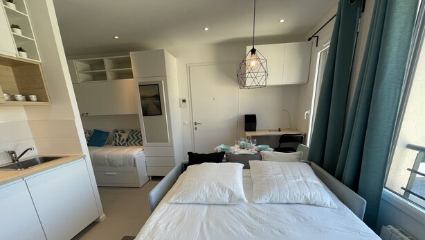 A modern hotel room with a large bed, a kitchenette, a dining area, and a view of the city.