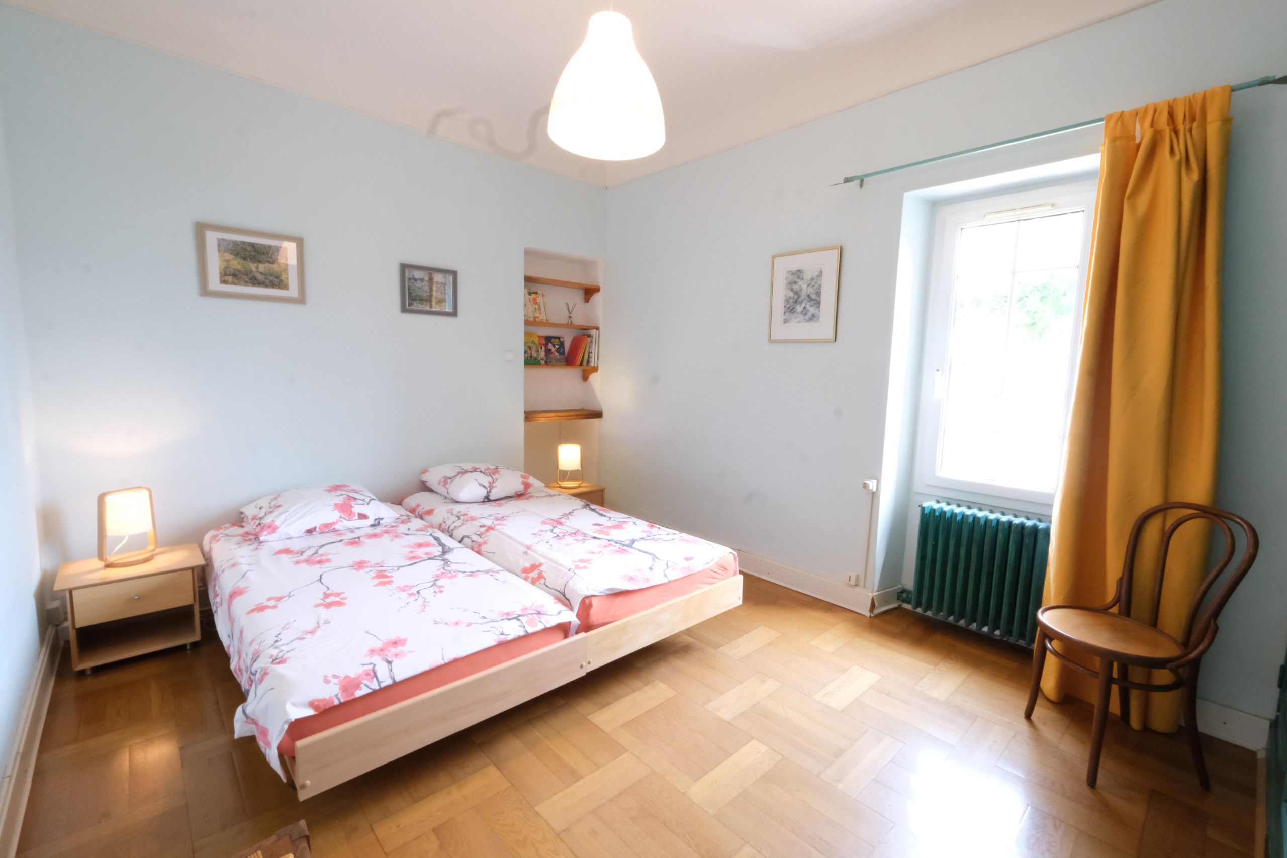 Une chambre à coucher comprenant un lit, une chaise, une table de chevet, une étagère avec des livres et une fenêtre avec des rideaux.