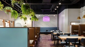 Un restaurant avec des banquettes, des tables, des plantes suspendues et un panneau néon représentant un hamburger.