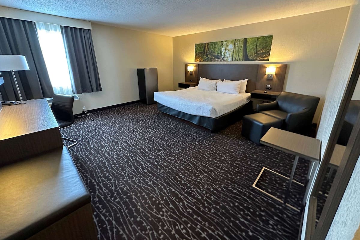 Ein Hotelzimmer mit einem großen Bett, einem Schreibtisch, einem Sessel, einem kleinen Tisch und einem großen Fenster mit Vorhängen.