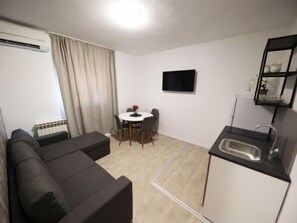 Ein kompakter Wohnbereich mit Sofa, Esstisch mit Stühlen, einer kleinen Küchenzeile mit Spüle und einem an der Wand befestigten Flachbildfernseher.