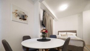 Une chambre d’hôtel avec une table de salle à manger ronde, quatre chaises, un lit avec une tête de lit, une table de chevet, un vase avec des fleurs et un tableau au mur.