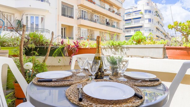 A table set for dining with plates, cutlery, and wine glasses on a balcony with a view of multi-story buildings and potted plants. - Apartment Hérold by Interhome (Nice)