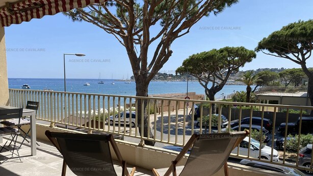 Un balcón con dos sillas plegables, una mesa y vistas al mar y coches aparcados.