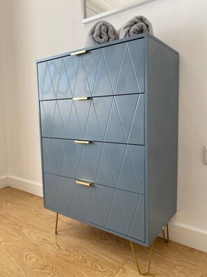 The cabinet features three drawers and a top shelf with rolled towels, supported by gold legs.
