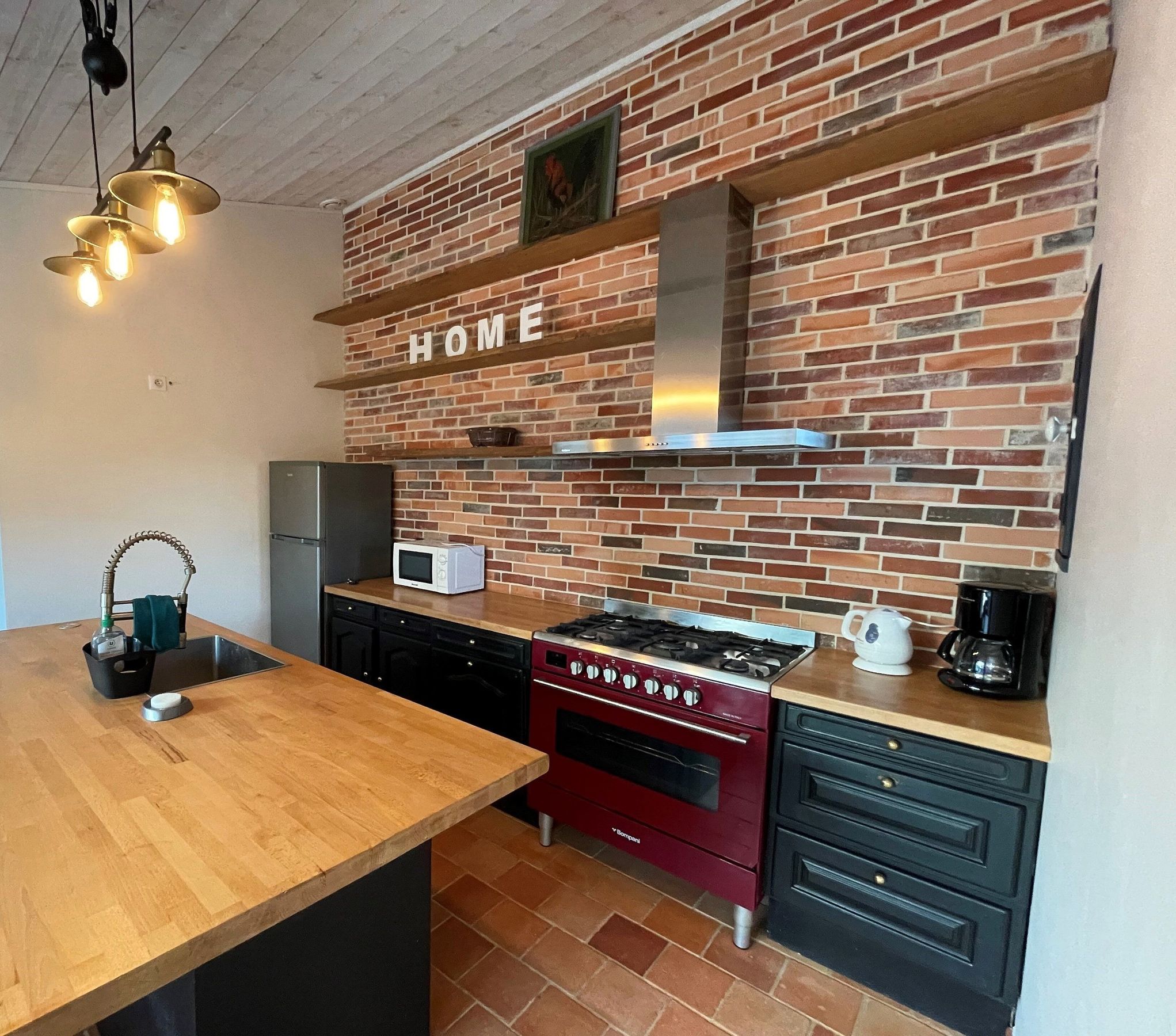 A kitchen with a brick wall, a wooden island, a stainless steel refrigerator, a microwave, a gas stove, and a coffee maker.