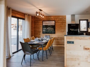 Un comedor moderno con una pared de madera, un ventanal grande y una cocina visible a través de una puerta abierta.