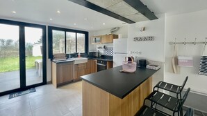 Una cocina moderna con una isla grande, armarios de madera y encimera negra. Hay una zona de comedor con sillas negras y una mesa pequeña. La cocina tiene un ventanal y una puerta que da a un patio.