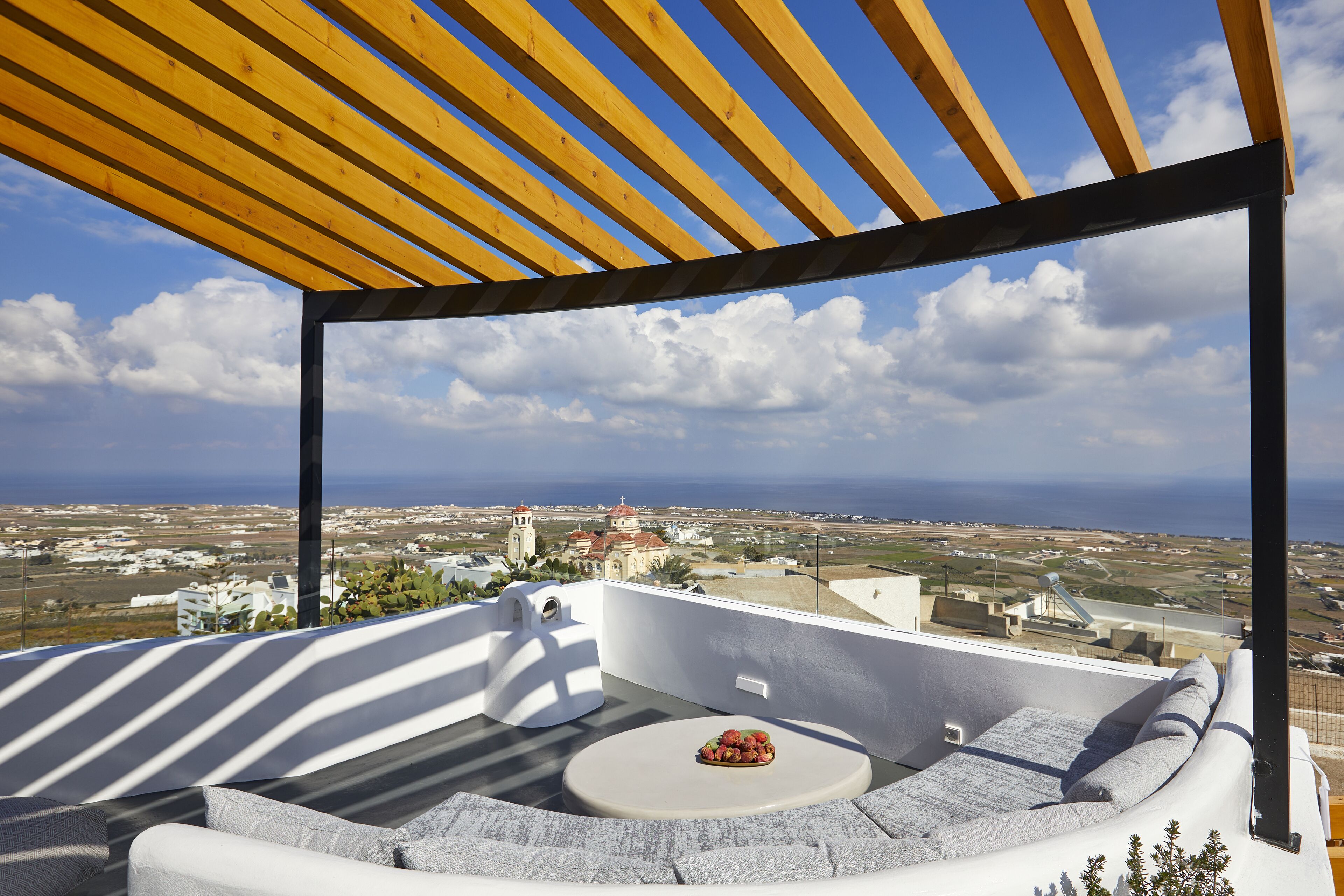 Una terraza en la azotea con una zona de estar con cojines blancos y una pequeña mesa con fruta, con vistas a un paisaje costero.