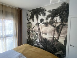 Un dormitorio con cama, un amplio mural tropical en la pared y una ventana con cortinas.