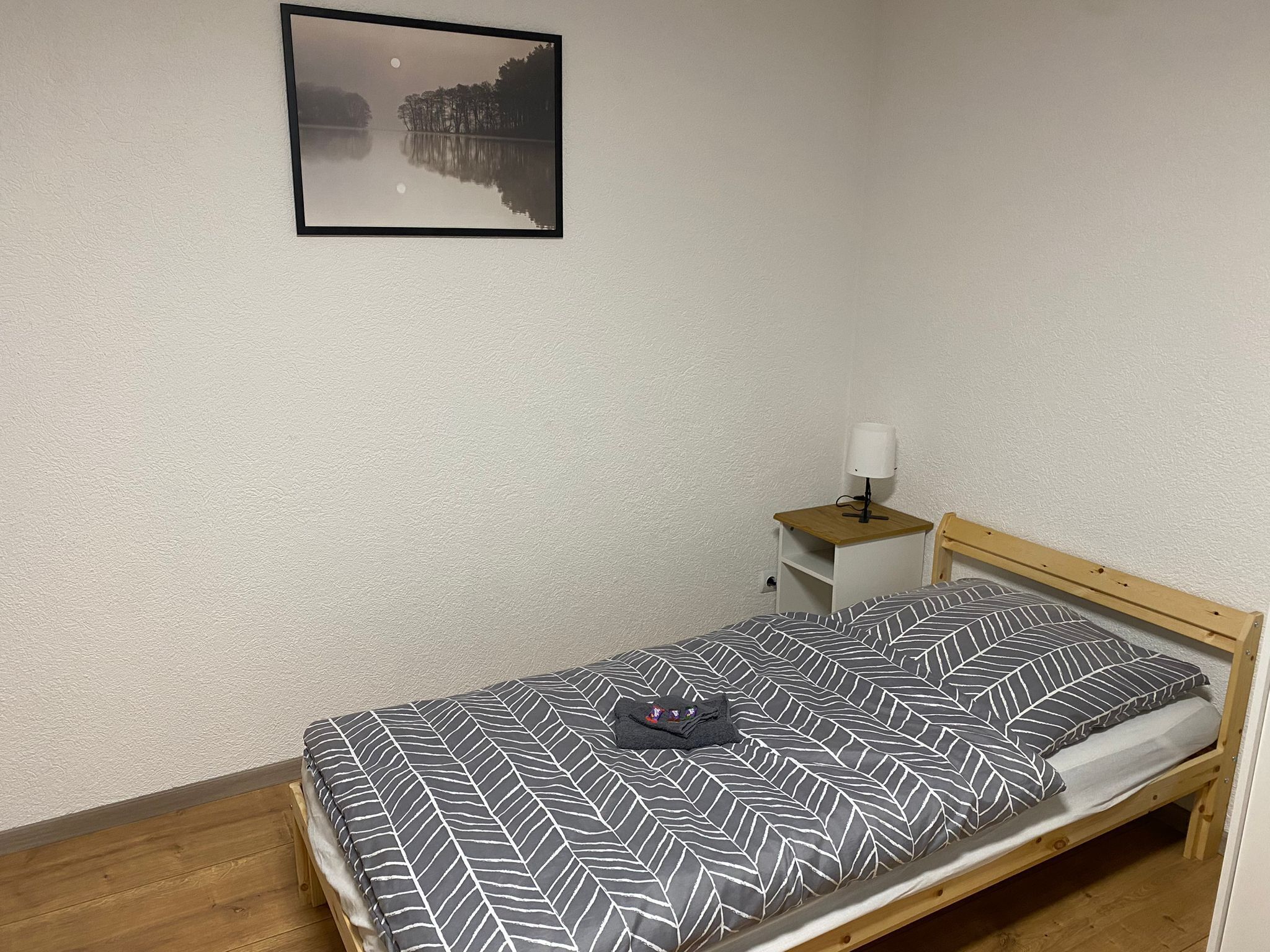 Ein Einzelbett mit gemustertem Bettbezug, ein Nachttisch mit Lampe und ein gerahmtes Bild an der Wand.