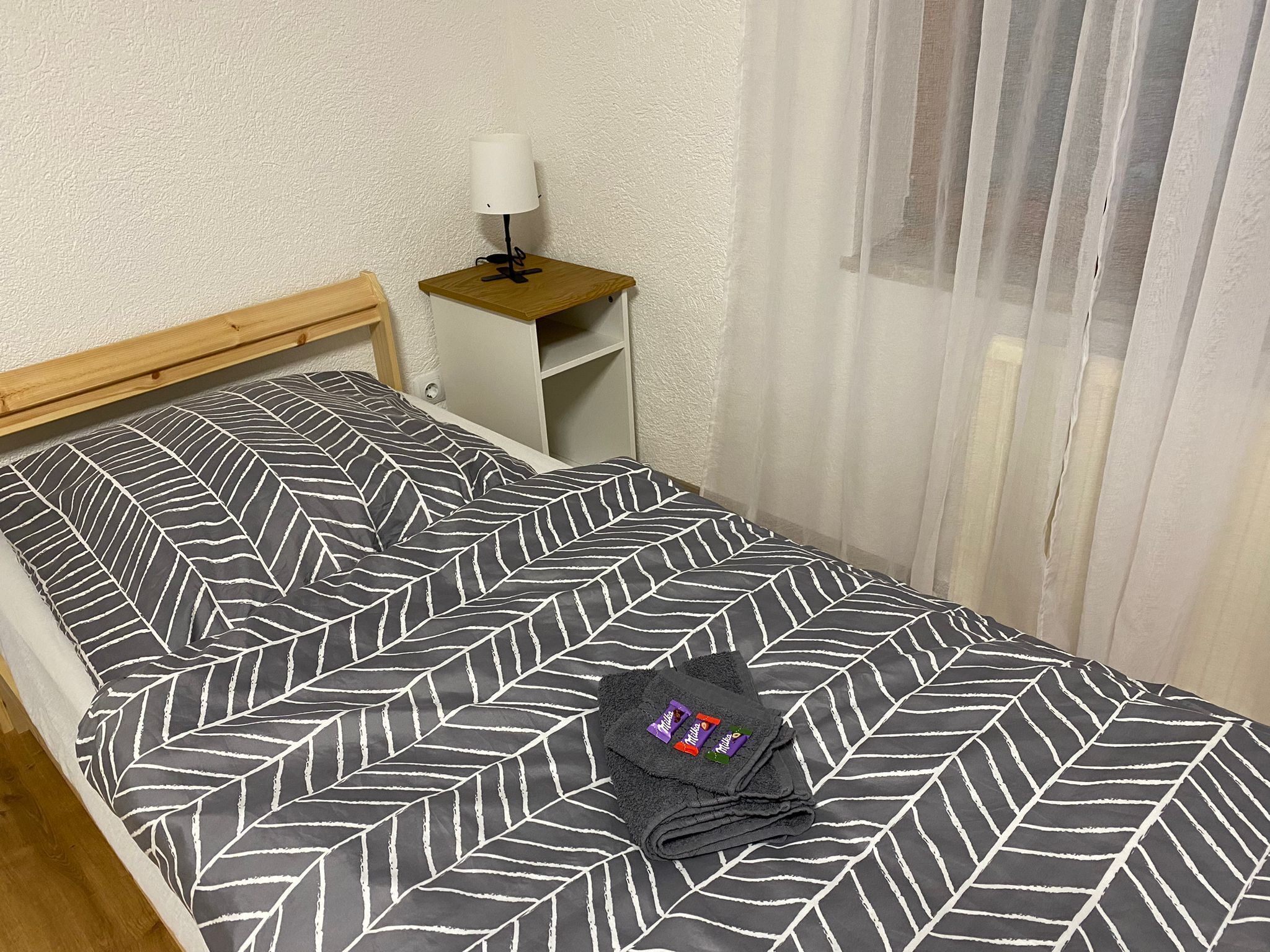 A neatly made bed with a geometric patterned bedspread, a small bedside table with a lamp, and a window with sheer curtains.