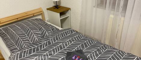 A neatly made bed with a geometric patterned bedspread, a small bedside table with a lamp, and a window with sheer curtains.